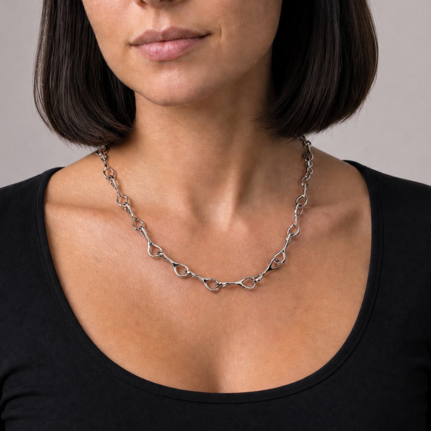 A close-up of a woman wearing a silver chain necklace with interlocking ring links, styled with a black scoop-neck top against a neutral gray background.