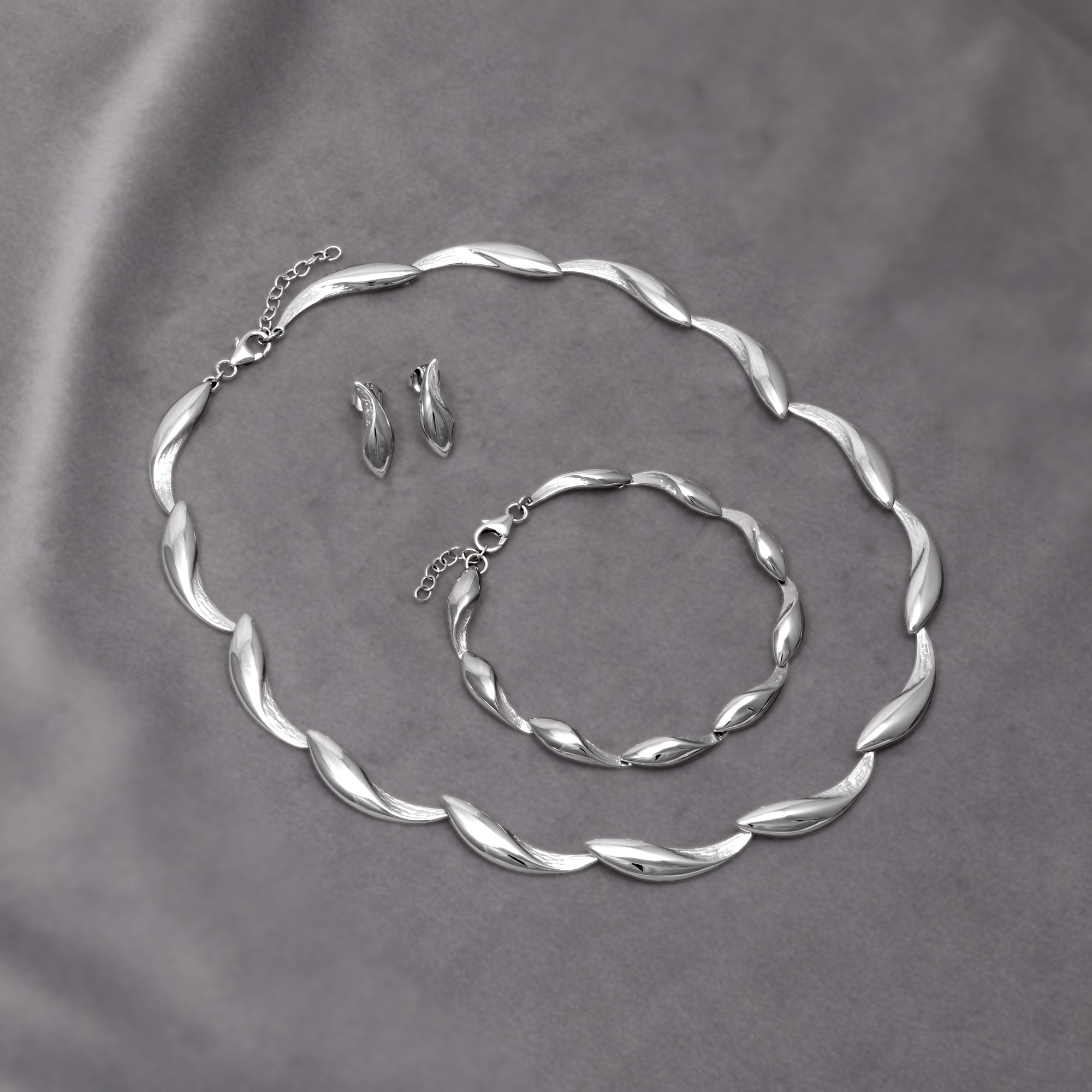 Set of sterling silver jewellery featuring a sculptural twisted wave motif, including a necklace, bracelet, and matching stud earrings with polished and textured finishes. Placed on a grey velvet cloth.