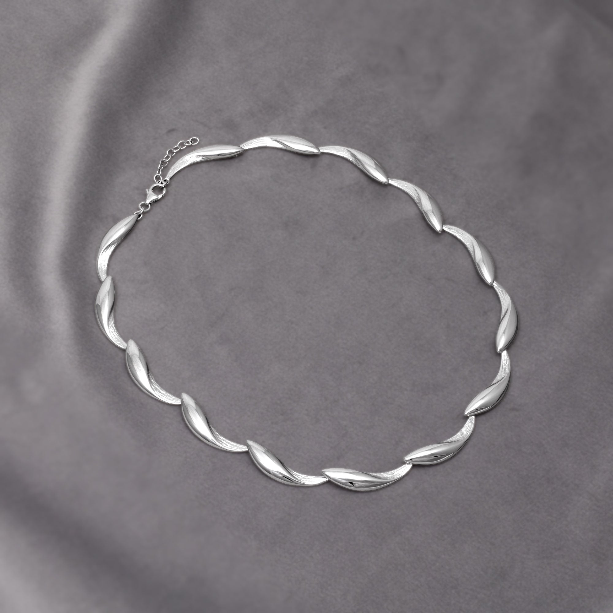 Silver jewellery featuring a sculptural twisted wave motif, a necklace with polished and textured finishes. Placed on a grey velvet cloth.
