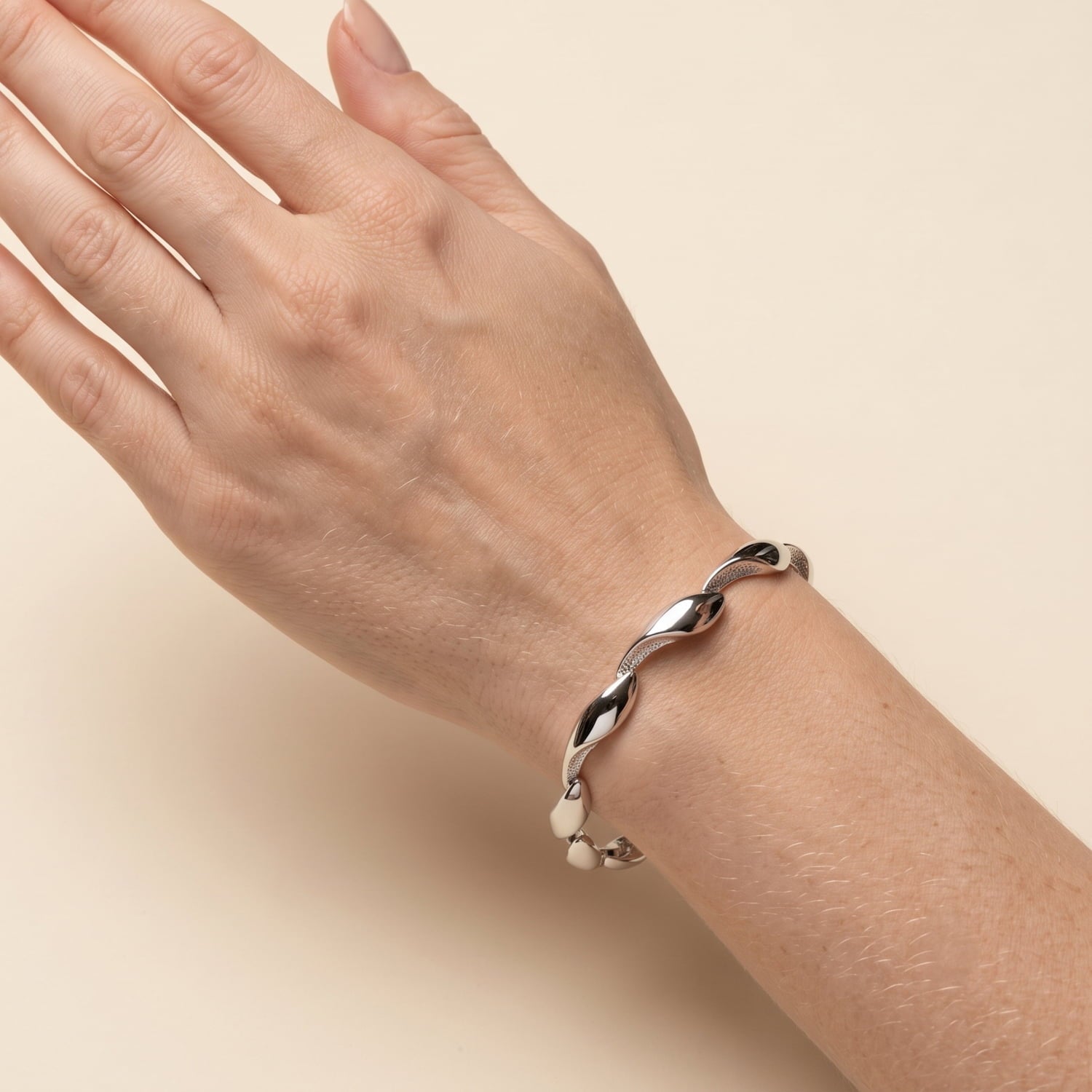 A close-up of a person's wrist wearing a polished silver twisted link bracelet against a solid beige background.