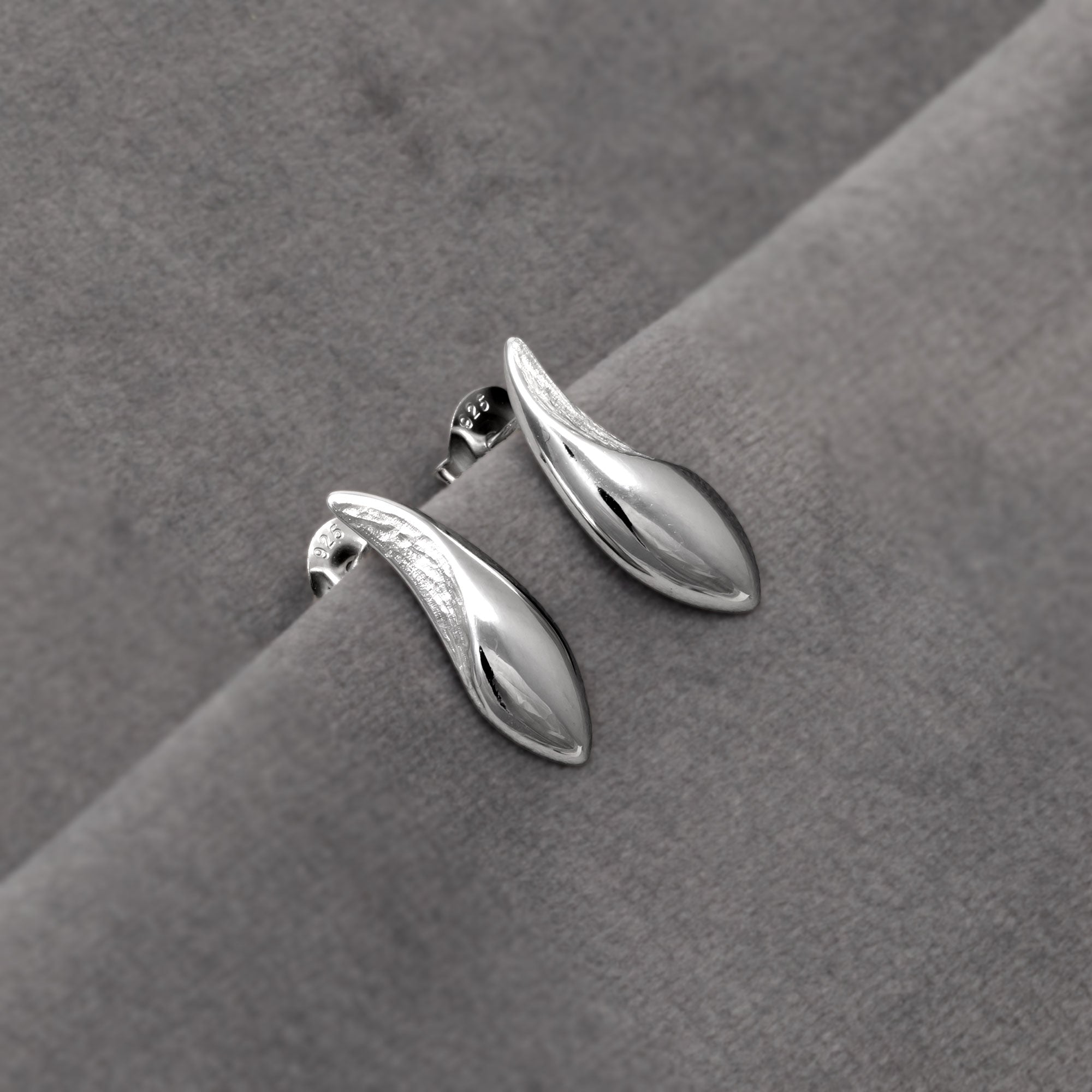 Polished and textured sterling silver stud earrings shaped as sleek, elongated droplets, featuring a smooth reflective surface with subtle sculptural detailing for modern elegance. Placed on a grey velvet cloth.