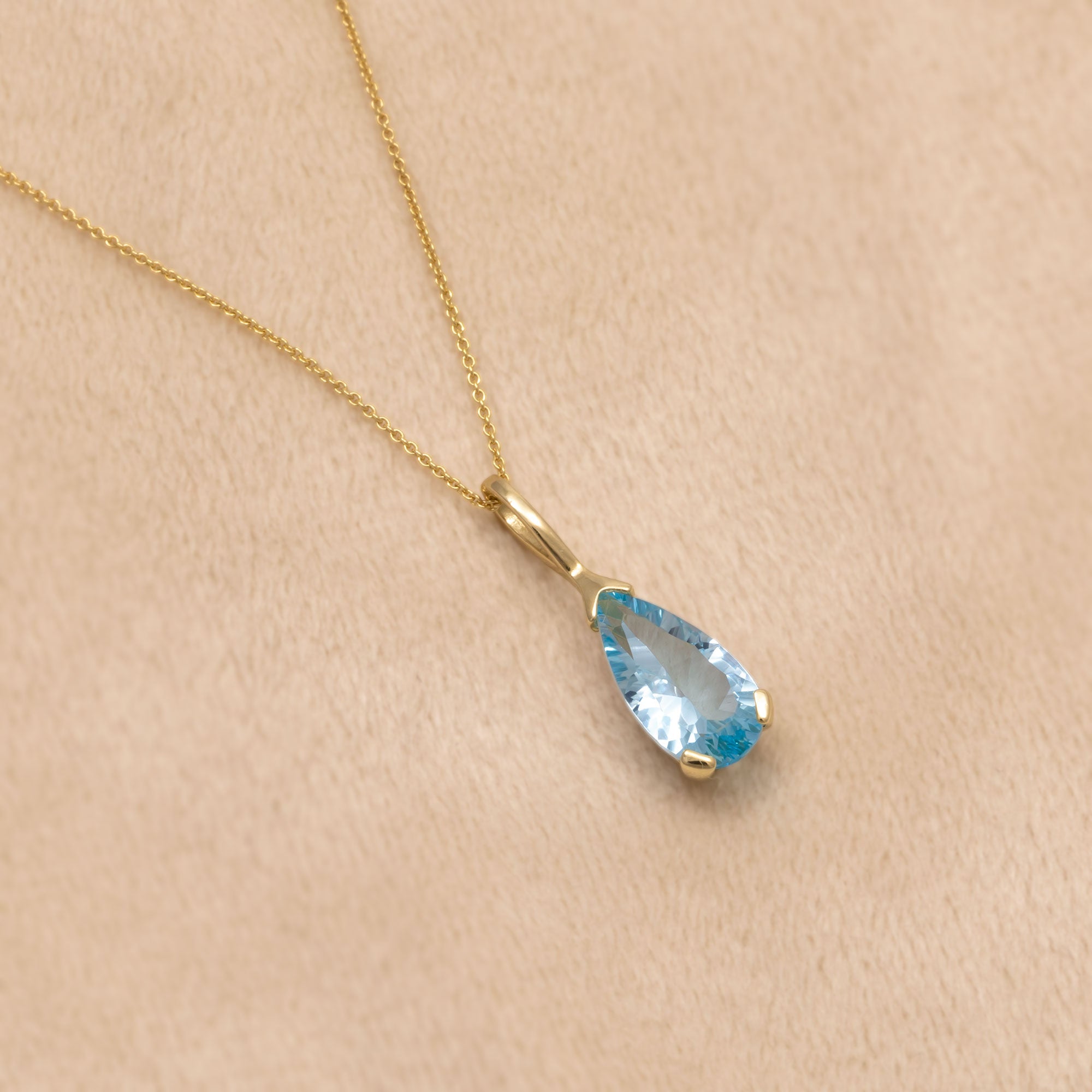 Elegant 9 carat gold necklace featuring a faceted blue topaz teardrop pendant suspended from a fine chain, showcasing a refined and contemporary design. Placed n a cream velvet cloth.
