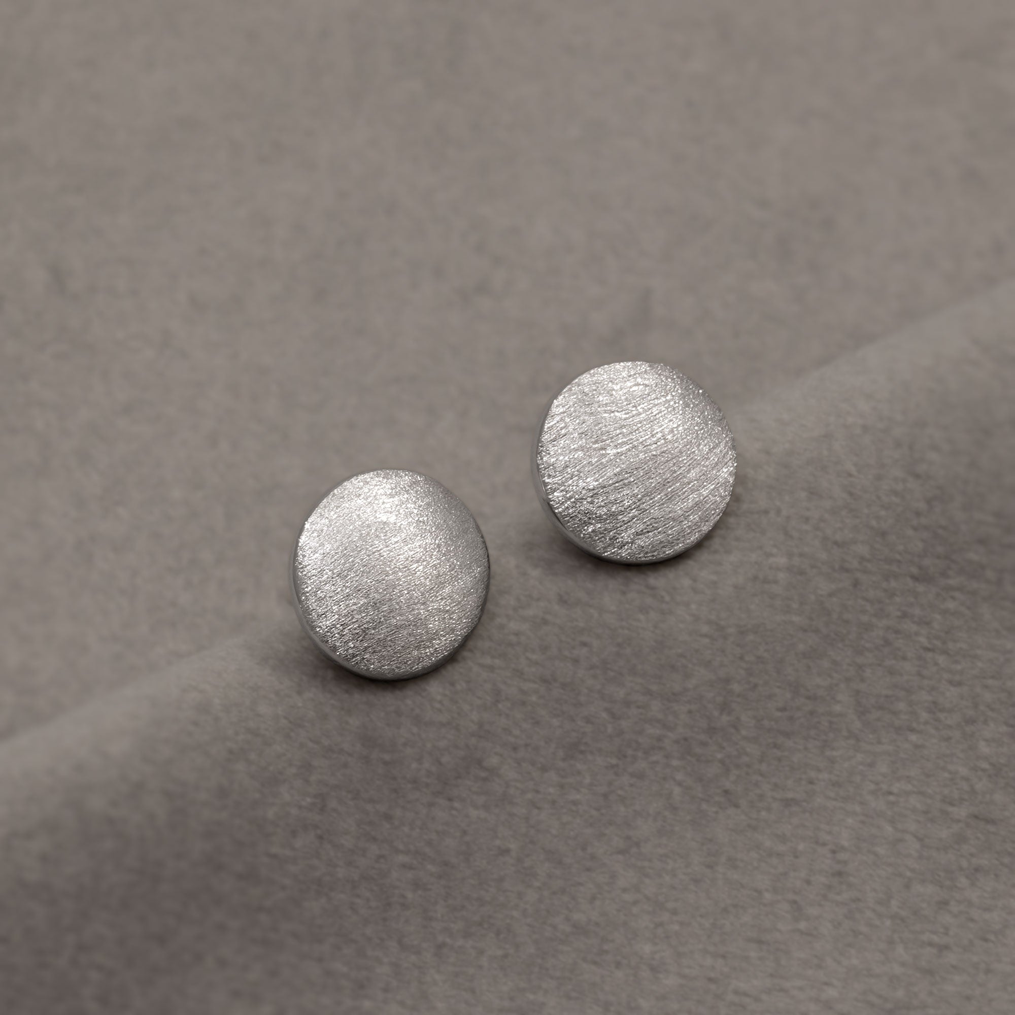 Pair of sterling silver stud earrings with a subtle satin finish, featuring a smooth, rounded design that highlights contemporary elegance and craftsmanship. Placed on a grey velvet cloth.