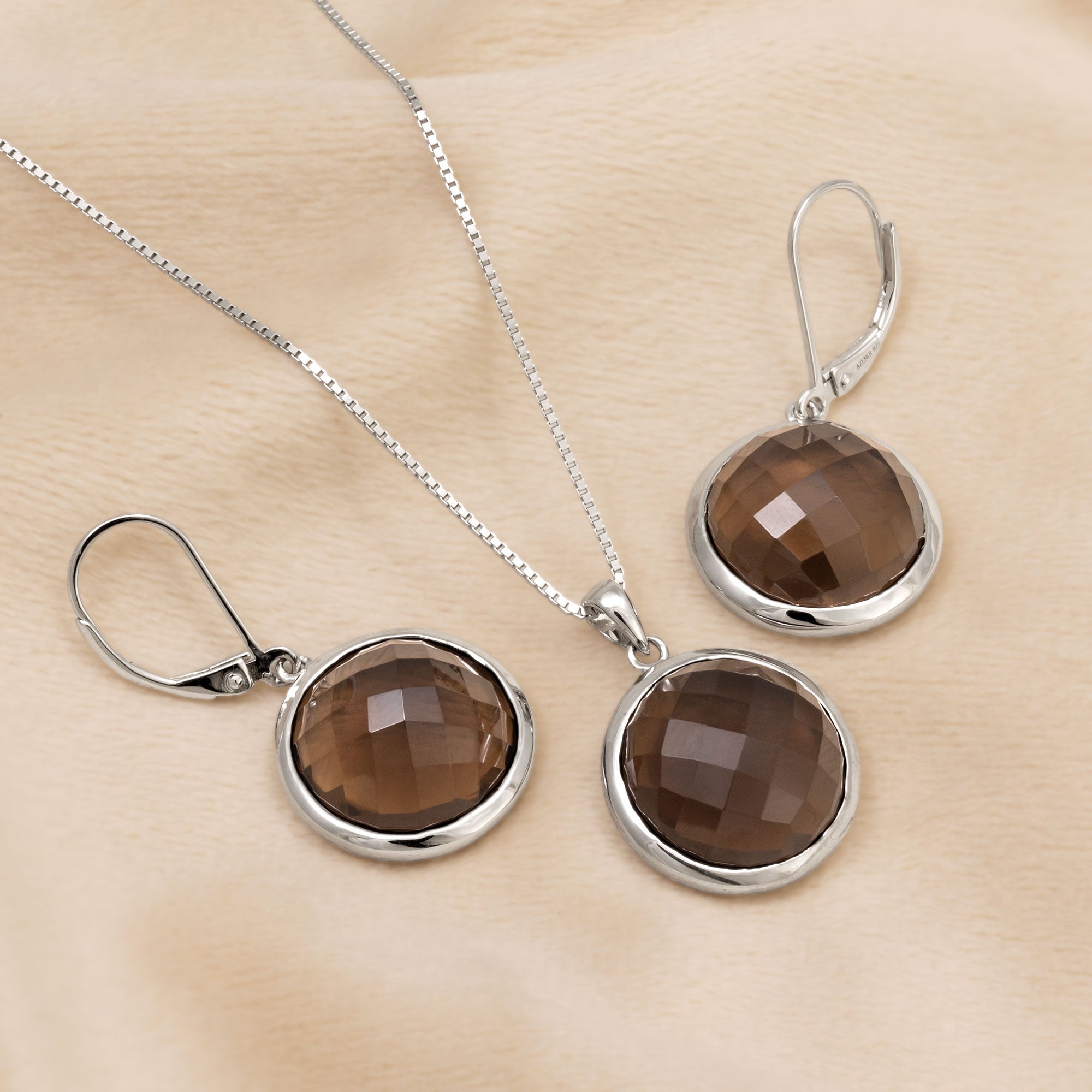 A sterling silver pendant and pair of drop earrings featuring round checker cut smoky quartz stones, designed with secure lever-back clasps for elegant wear. Placed on a cream velvet cloth.