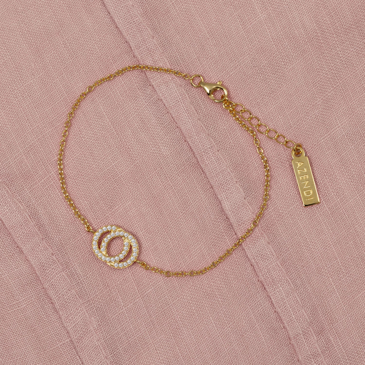 Delicate gold vermeil bracelet featuring interlocking circles embellished with sparkling cubic zirconia, adjustable chain, and a branded Azendi tag on a soft pink fabric background.