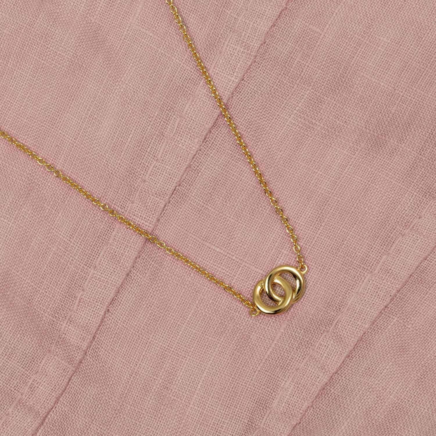 Yellow gold vermeil necklace featuring two interlocking circles on an adjustable chain, presented on a soft textured pink fabric background.