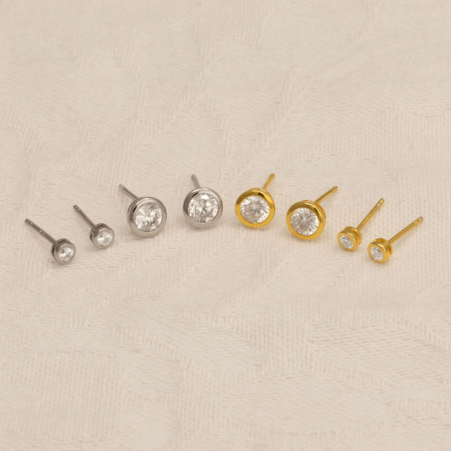 Collection of gold vermeil and sterling silver stud earrings with brilliant cut clear stones in varying sizes, arranged on a soft beige textured fabric background.
