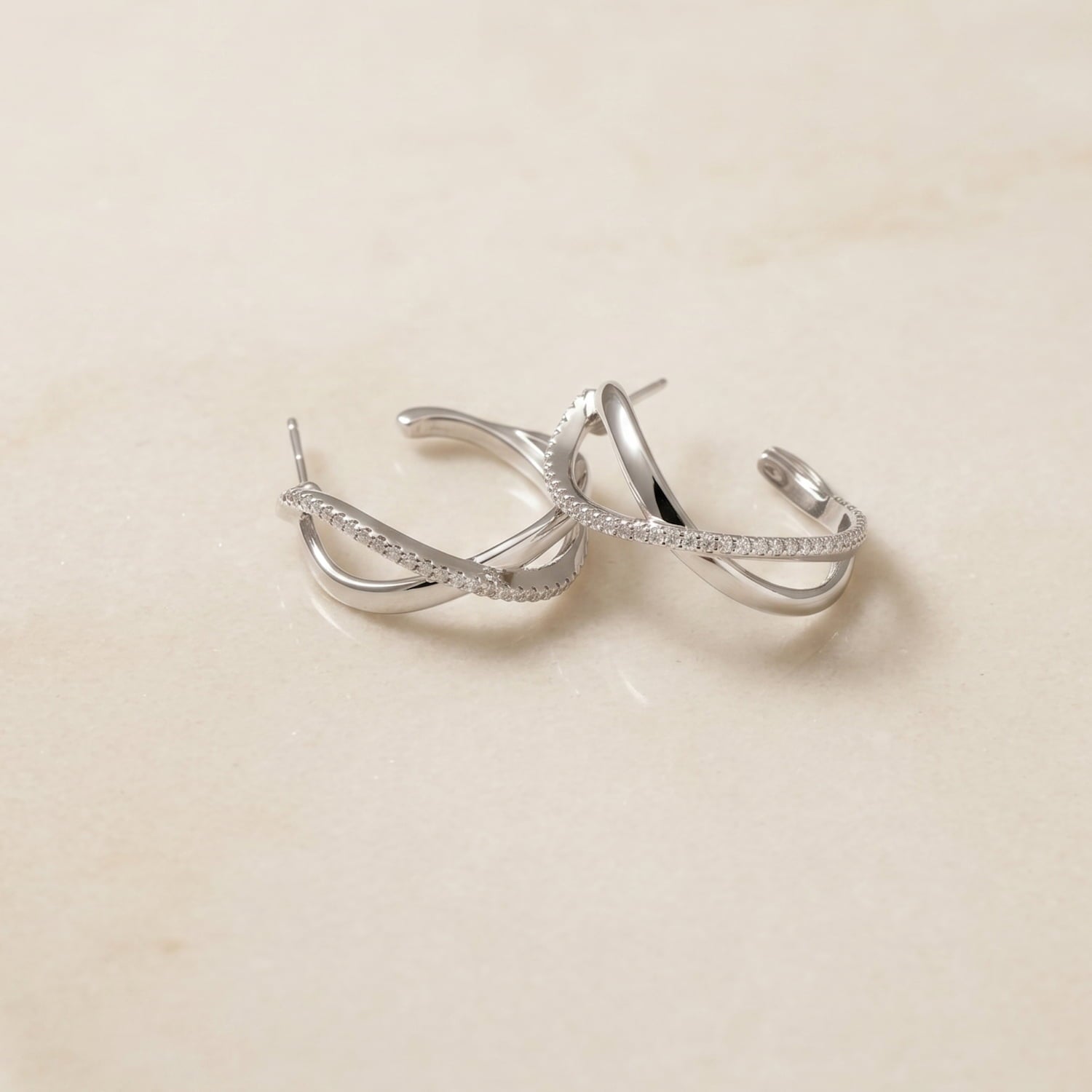 Pair of large sterling silver hoop earrings featuring an elegant crossover wave design accented with pavé-set stones, laid on a marble background.