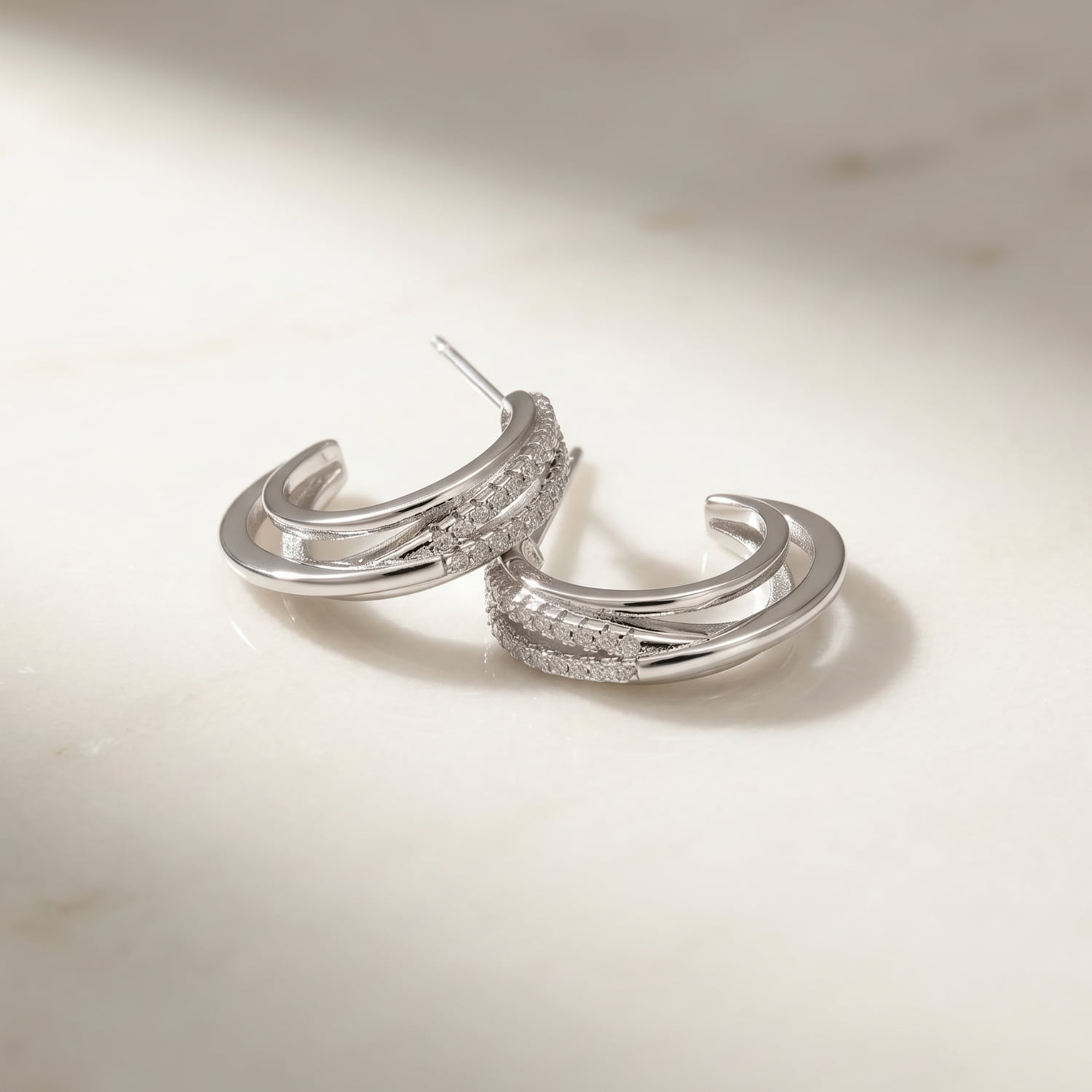 A pair of sterling silver hoop earrings featuring a four-strand crossover design with pavé-set stones laid upon a marble background.