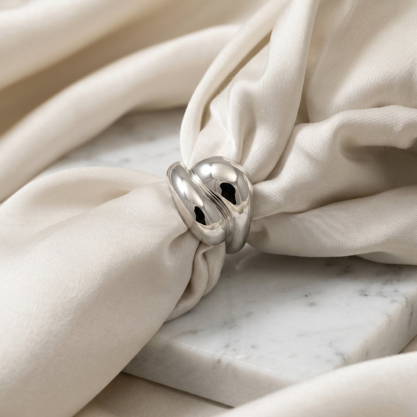 Close-up of a curving, double teardrop silver ring on cream fabric with a marble surface background.