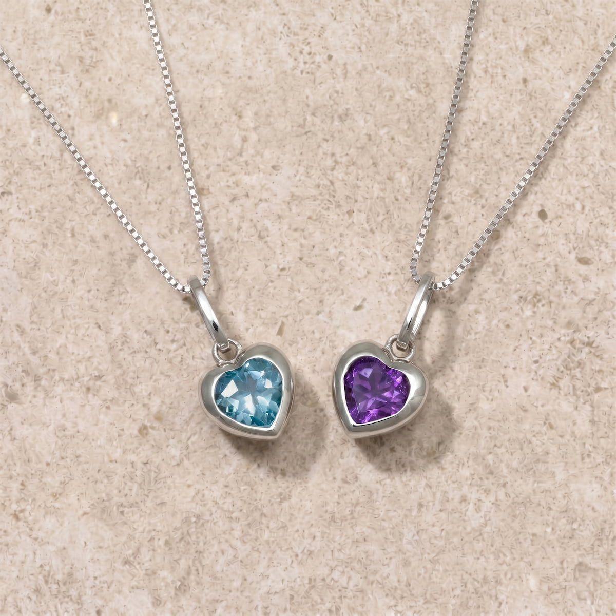 Two heart-shaped pendants with blue topaz and amethyst on silver chains on a beige surface
