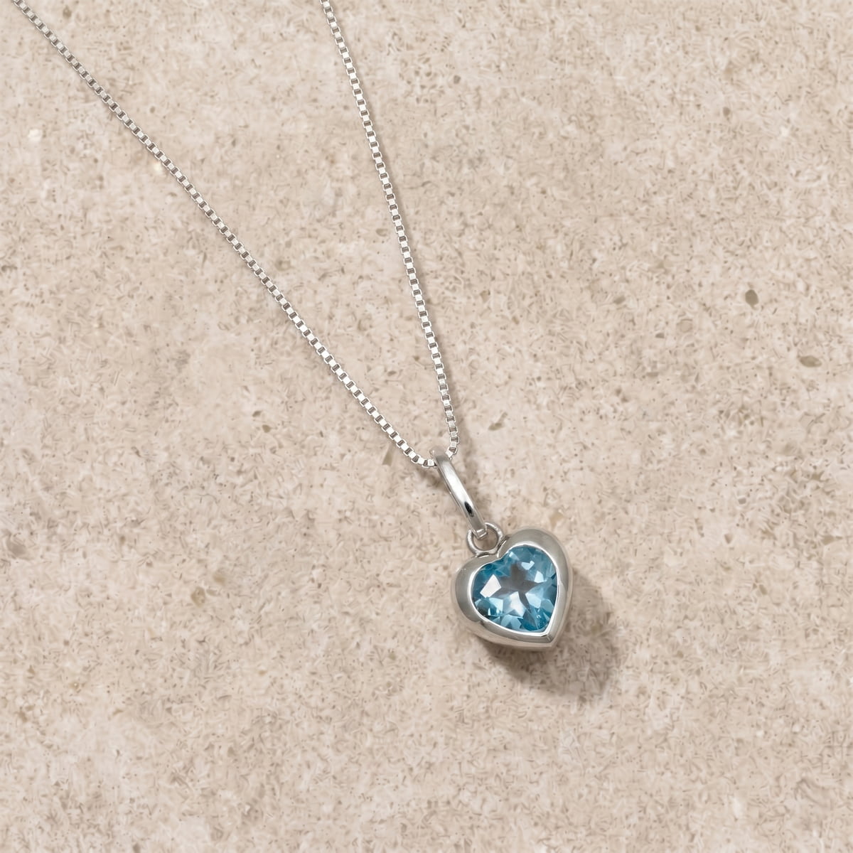 Silver and heart-shaped pendant with blue topaz gemstone on a beige limestone surface