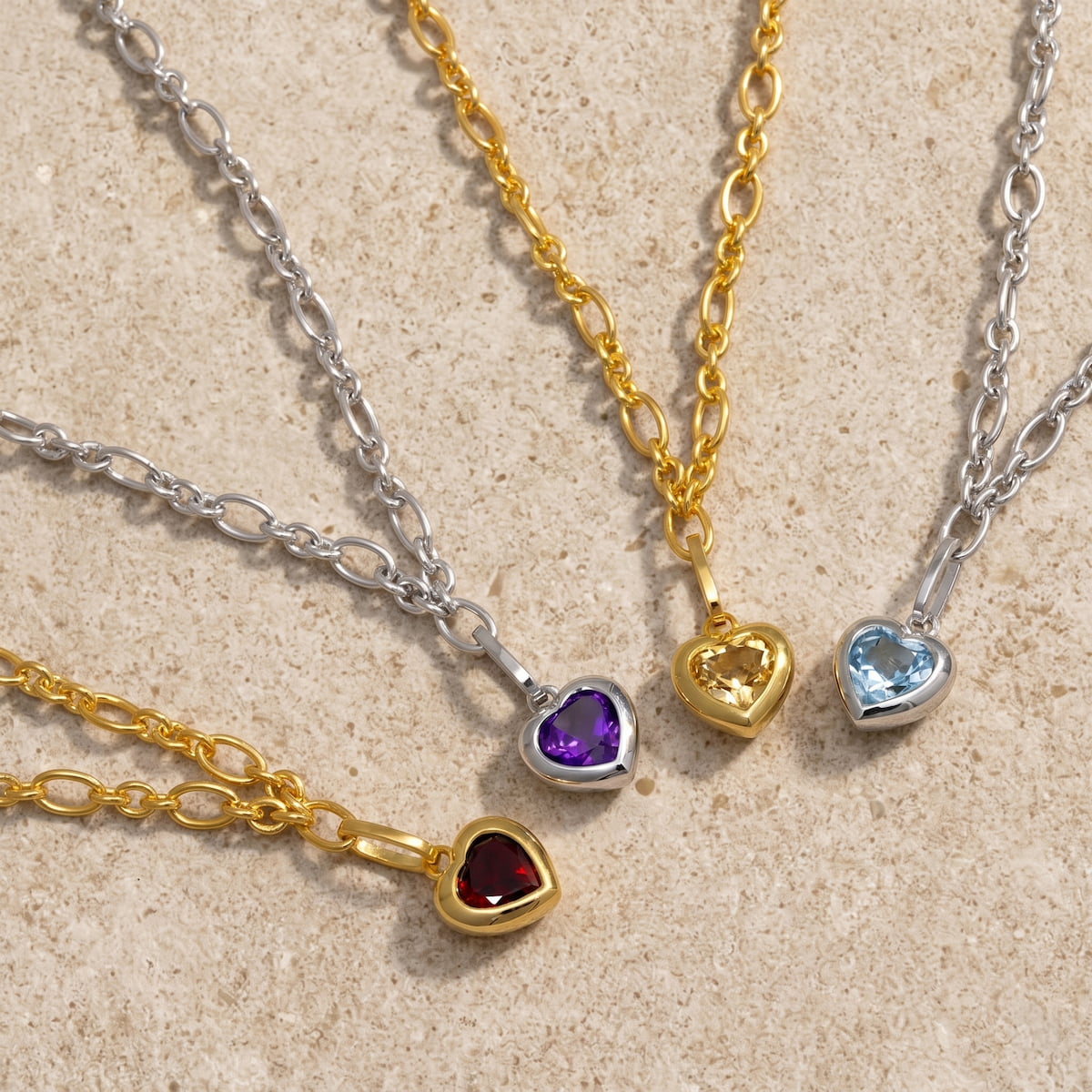 Four heart-shaped charm bracelets on gold vermeil and silver chains against a beige stone background