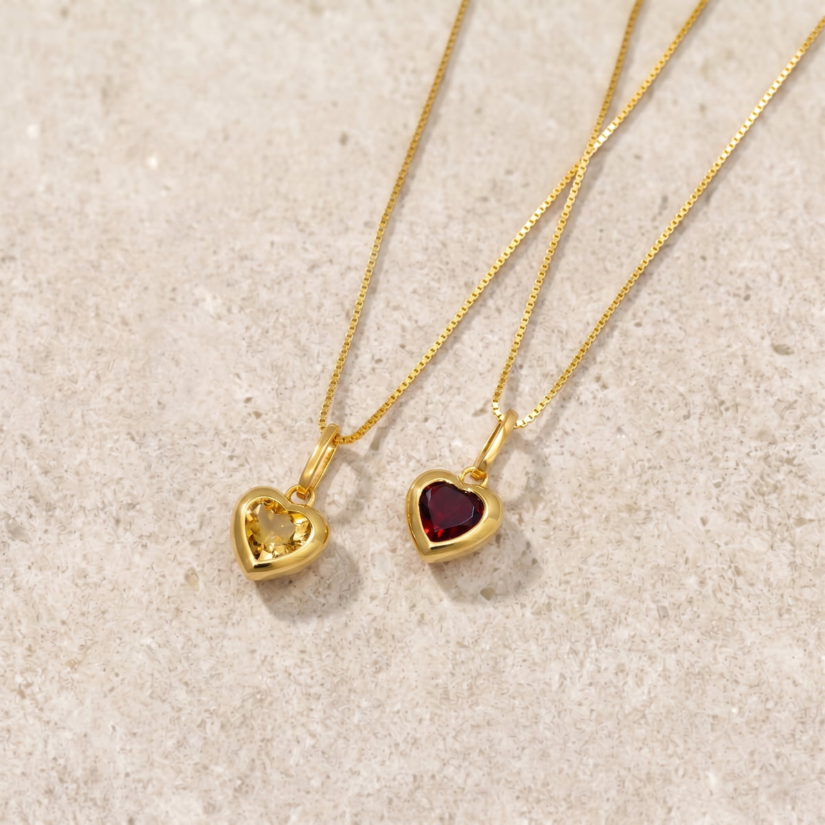 Two gold vermeil heart-shaped pendants with citrene and garnet gemstones on a beige limestone surface