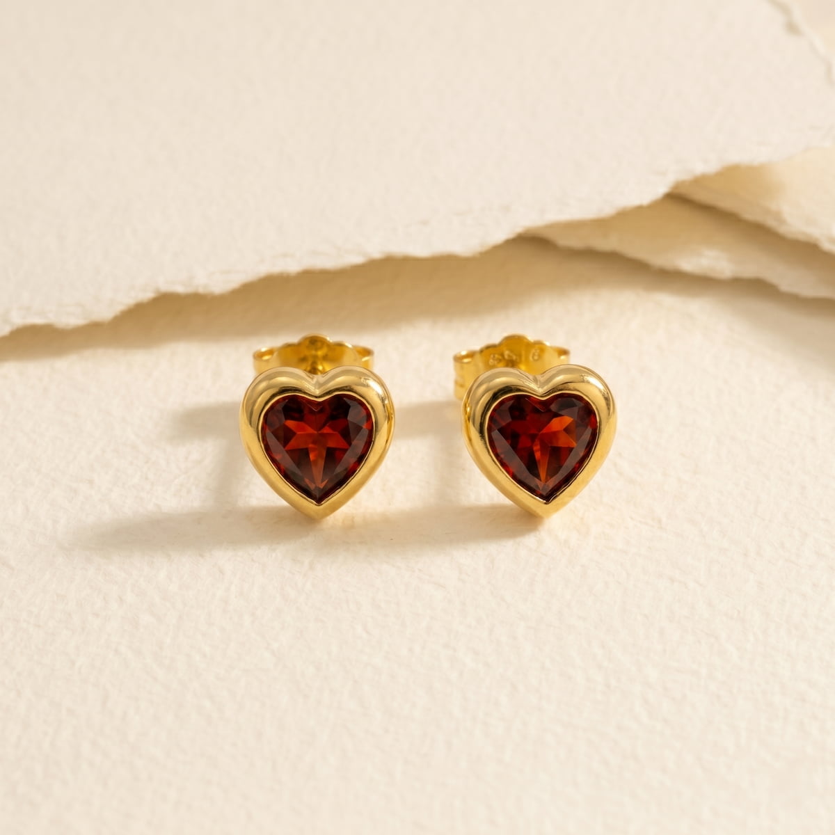 Heart-shaped gold vermeil earrings with red garnet gemstones on a paper background