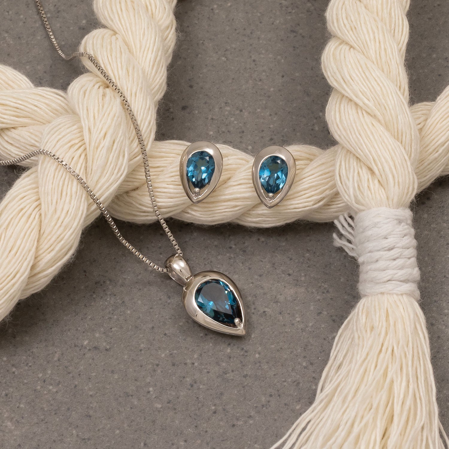 Pair of sterling silver stud earrings set with deep blue teardrop-shaped London Blue Topaz gemstones, displayed alongside a matching pendant on a delicate silver chain.