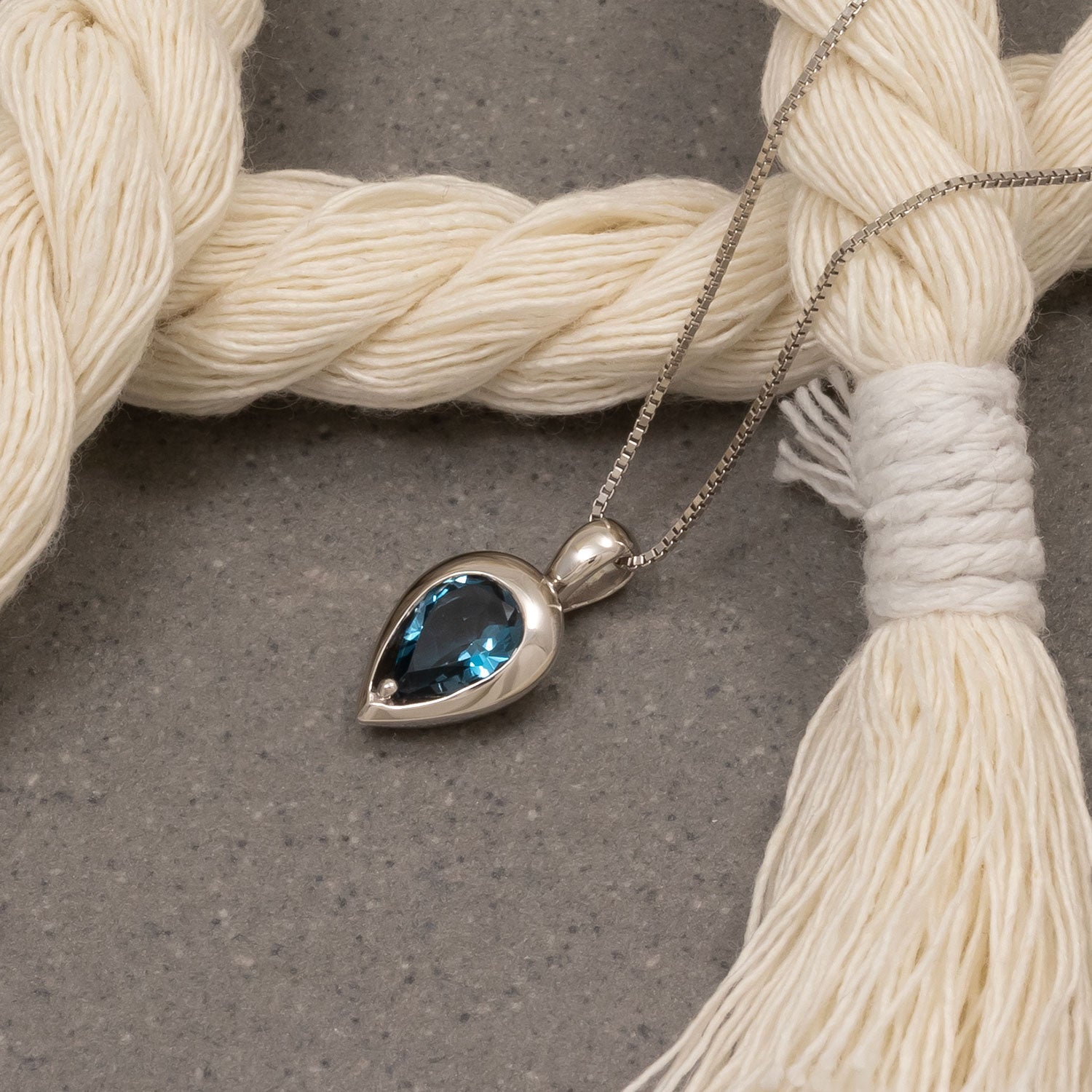 Teardrop-shaped sterling silver pendant featuring a deep London blue topaz gemstone, suspended from a delicate silver chain, photographed against neutral textured yarn.