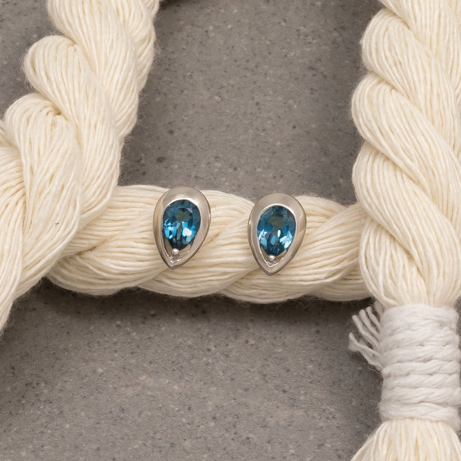 Pair of sterling silver stud earrings set with London Blue Topaz gemstones in a teardrop shape, presented on a neutral textured background with natural-fiber rope accents.