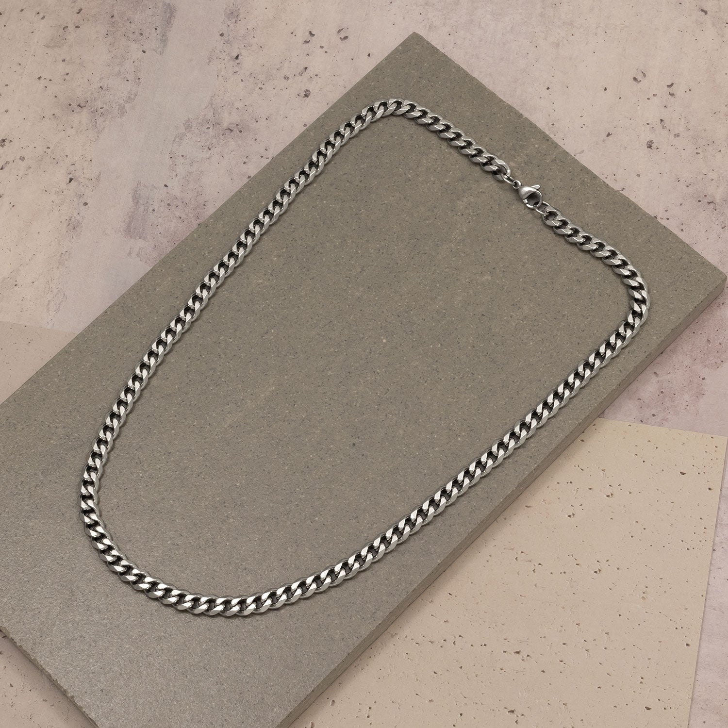 Sterling silver men's curb chain necklace with a polished finish, displayed on layered neutral-toned textured surfaces highlighting its bold, sculptural design.
