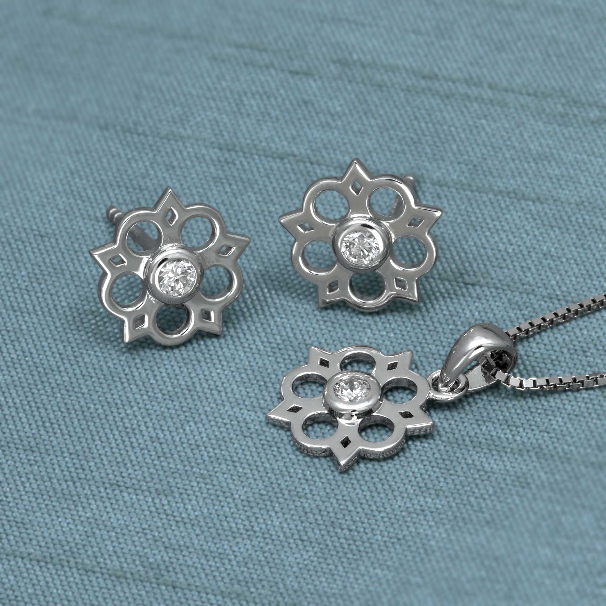 Sterling silver Yorkshire rose pendant featuring a 0.07 carat lab-grown diamond at the centre, suspended on a delicate fine chain with sculptural elegance. Delicate sterling silver stud earrings featuring an openwork Yorkshire rose motif with a central lab-grown diamond, combining elegant floral design with contemporary silver jewellery. Placed on a teal satin cloth.