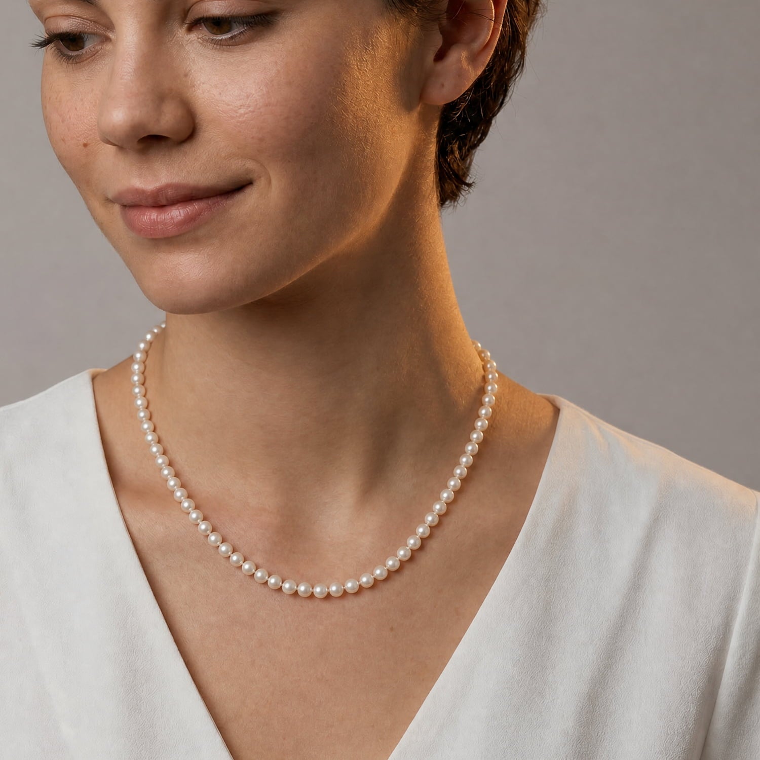 A close-up of a woman wearing a classic single-strand pearl necklace with white pearls and a white V-neck top, set against a neutral gray background.