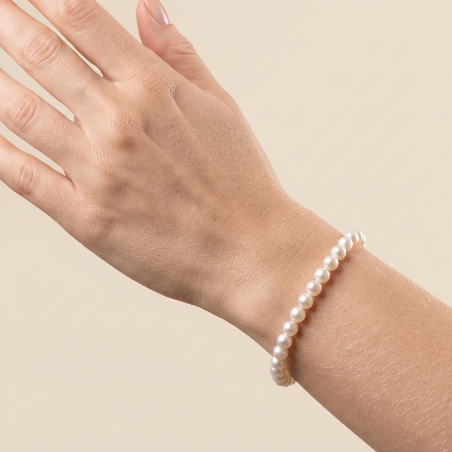 A close-up of a person's wrist wearing a delicate strand bracelet made of small, round, lustrous white pearls, set against a solid, warm beige background.
