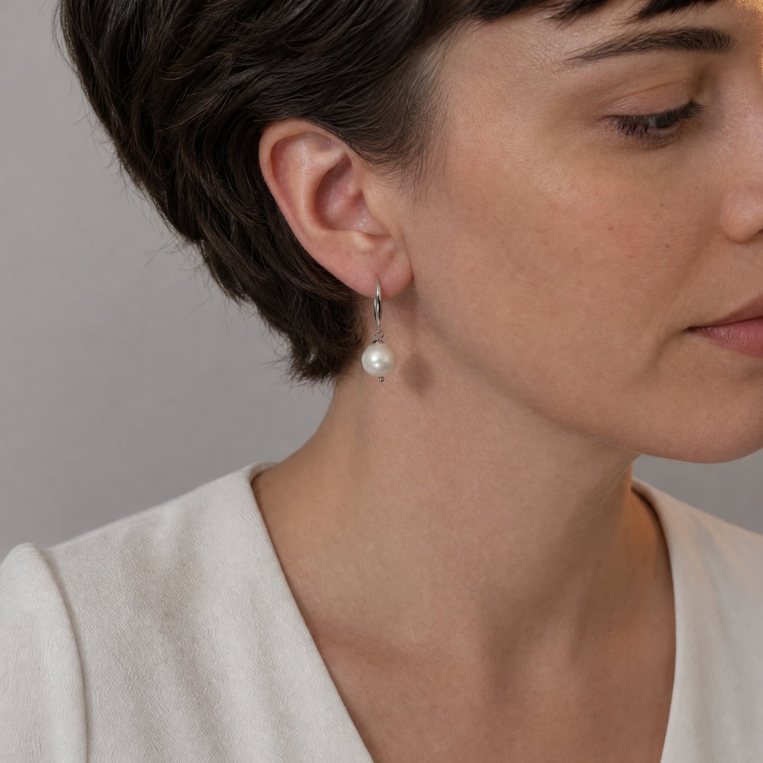 A close-up side profile of a woman with short dark hair wearing a delicate silver drop earring with a single white pearl, against a neutral gray background.