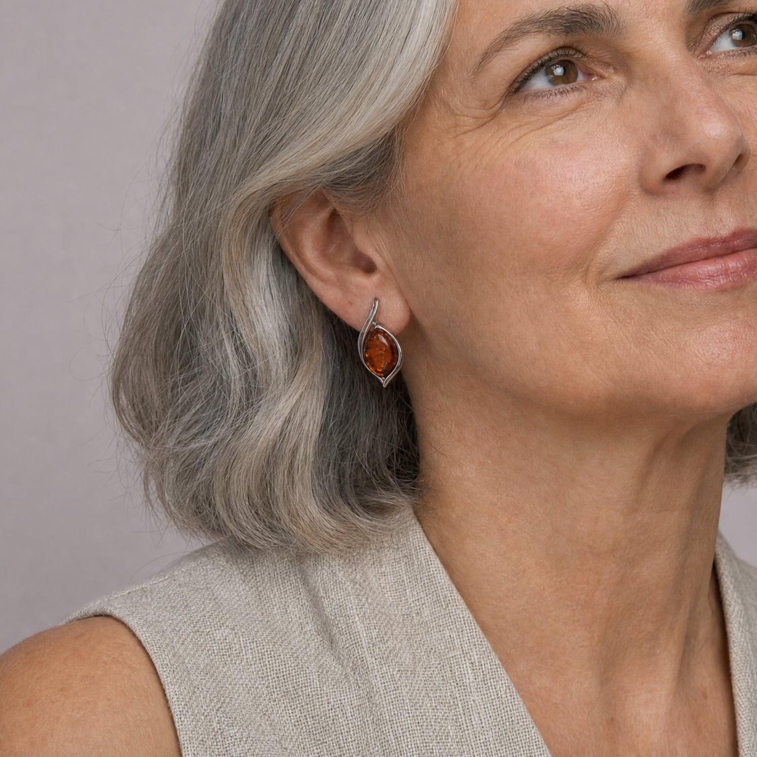 A close-up of a woman with grey hair wearing a silver teardrop earring with a warm amber stone, set against a neutral gray background.