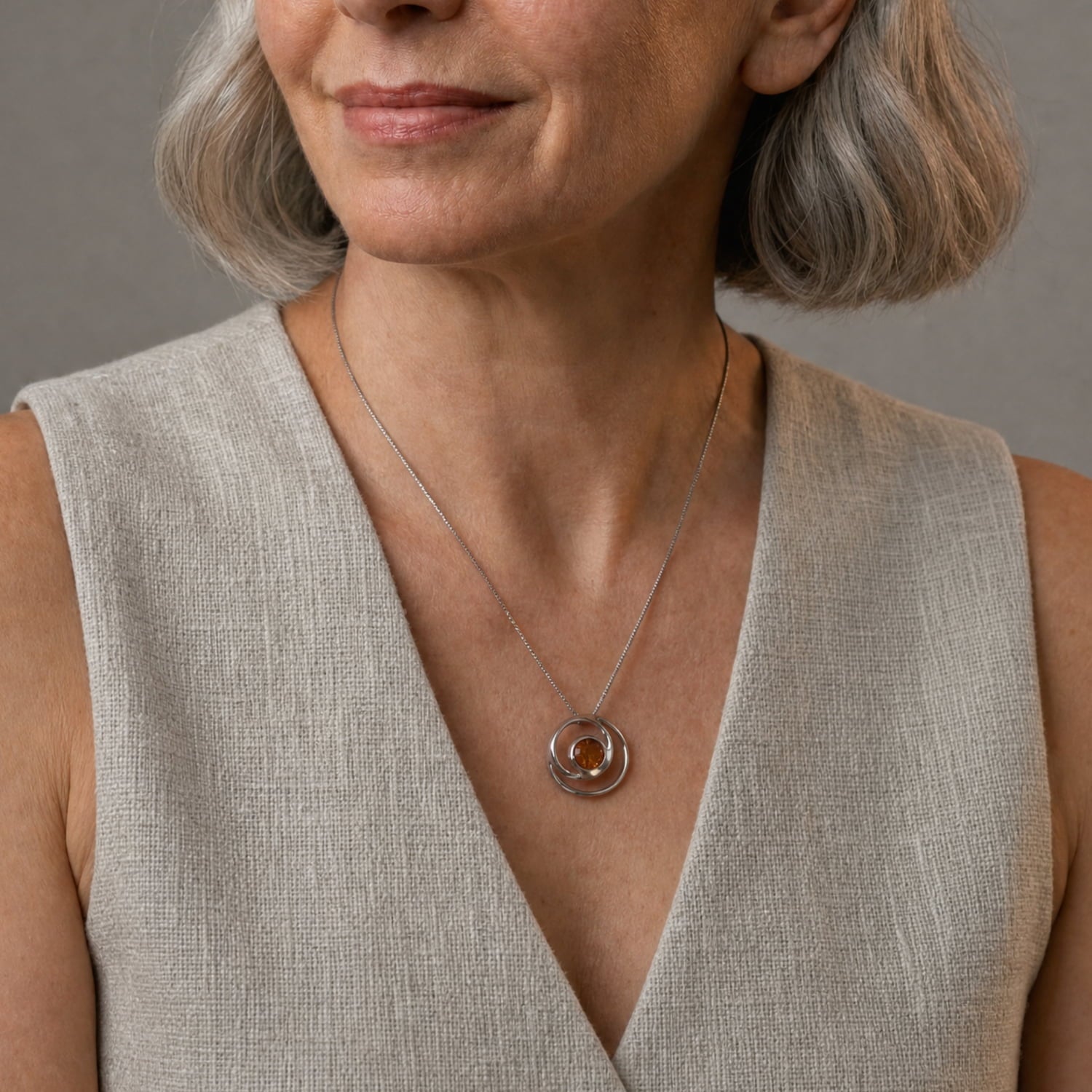 A close-up of a woman wearing a silver circular pendant necklace with a warm amber center, paired with a textured beige sleeveless top against a neutral gray background.