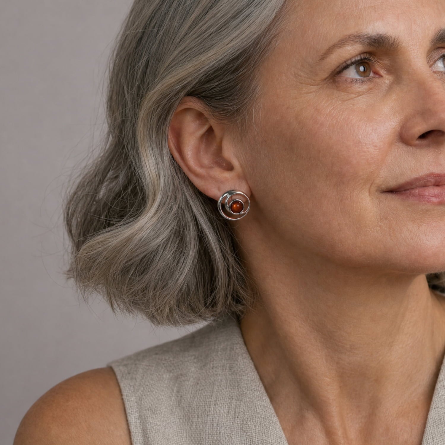 A close-up of a woman with grey hair wearing a silver circle earring with an amber gem, against a neutral gray background.
