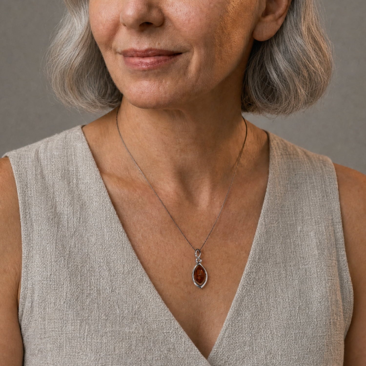 A close-up of a woman wearing a silver twist pendant with an amber gem, paired with a beige linen top against a neutral gray background.