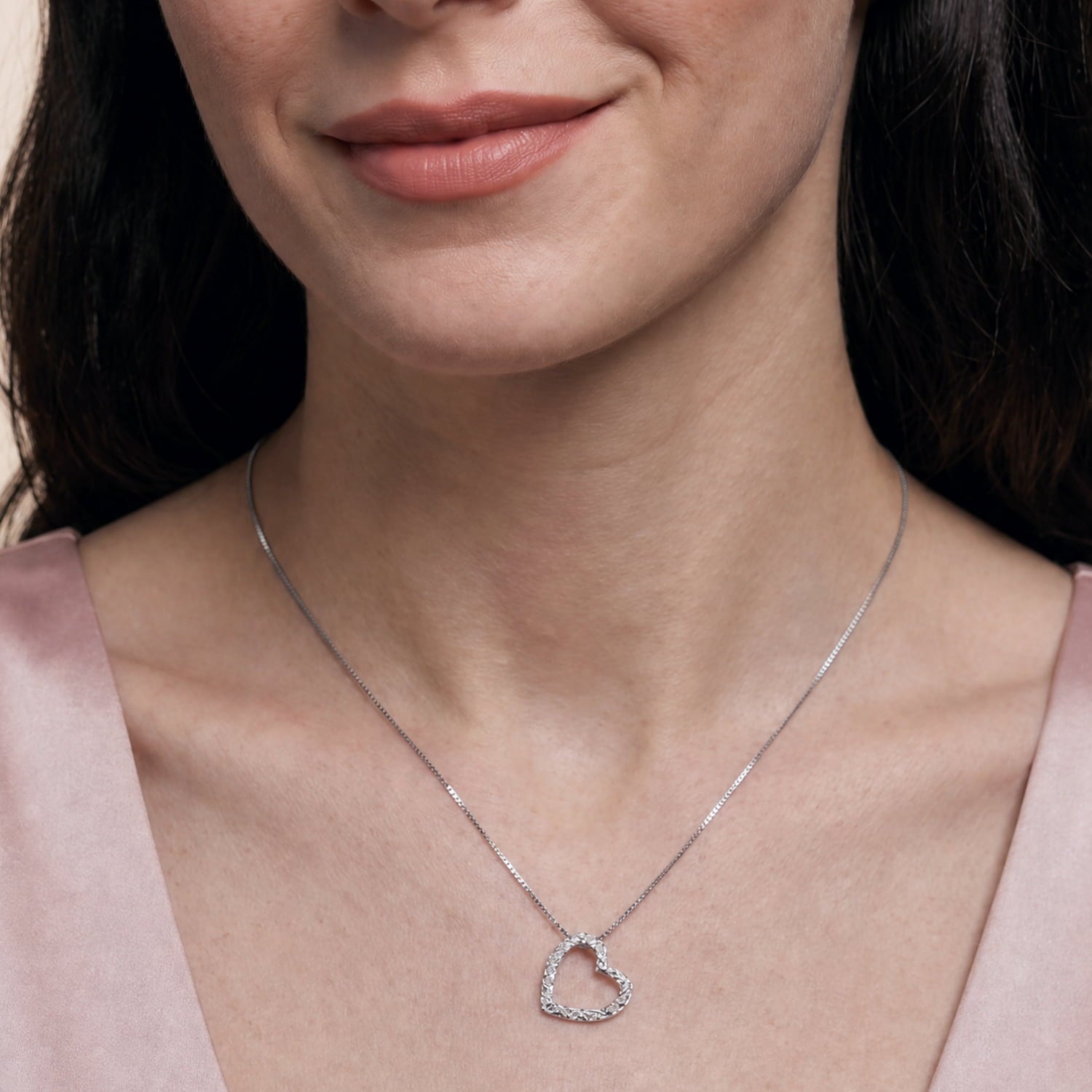A close-up of a woman wearing a delicate silver necklace with a heart-shaped pendant featuring a textured finish, paired with a soft pink satin top.