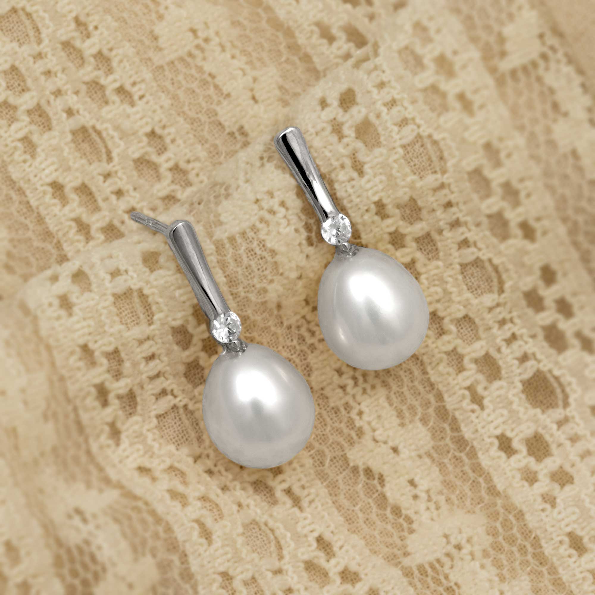 Sterling silver drop earrings featuring large teardrop freshwater pearls accented with sparkling cubic zirconia stones, displayed on a soft lace fabric background.