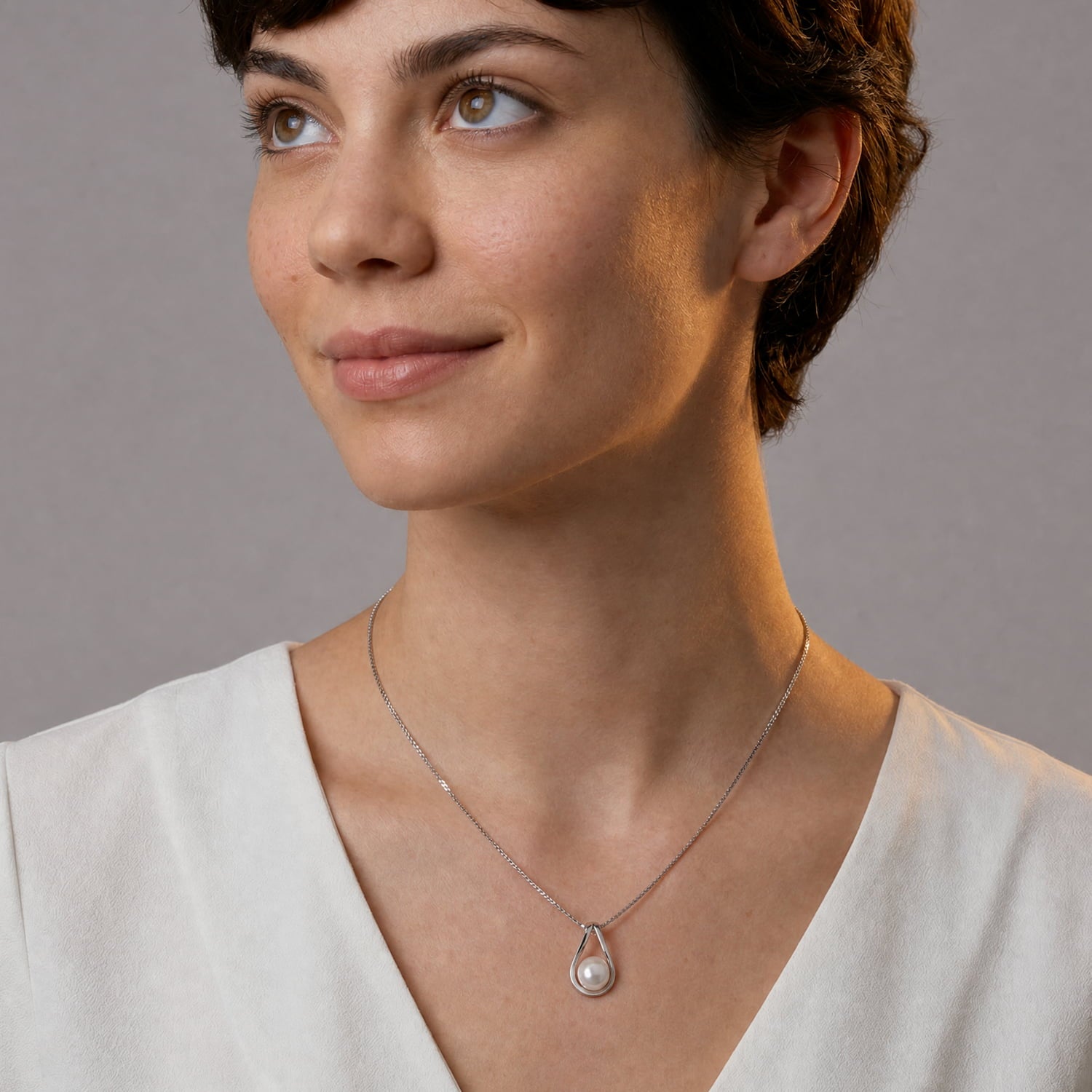 A close-up of a woman wearing a delicate silver chain necklace with a teardrop-shaped white pearl pendant, set against a neutral gray background.