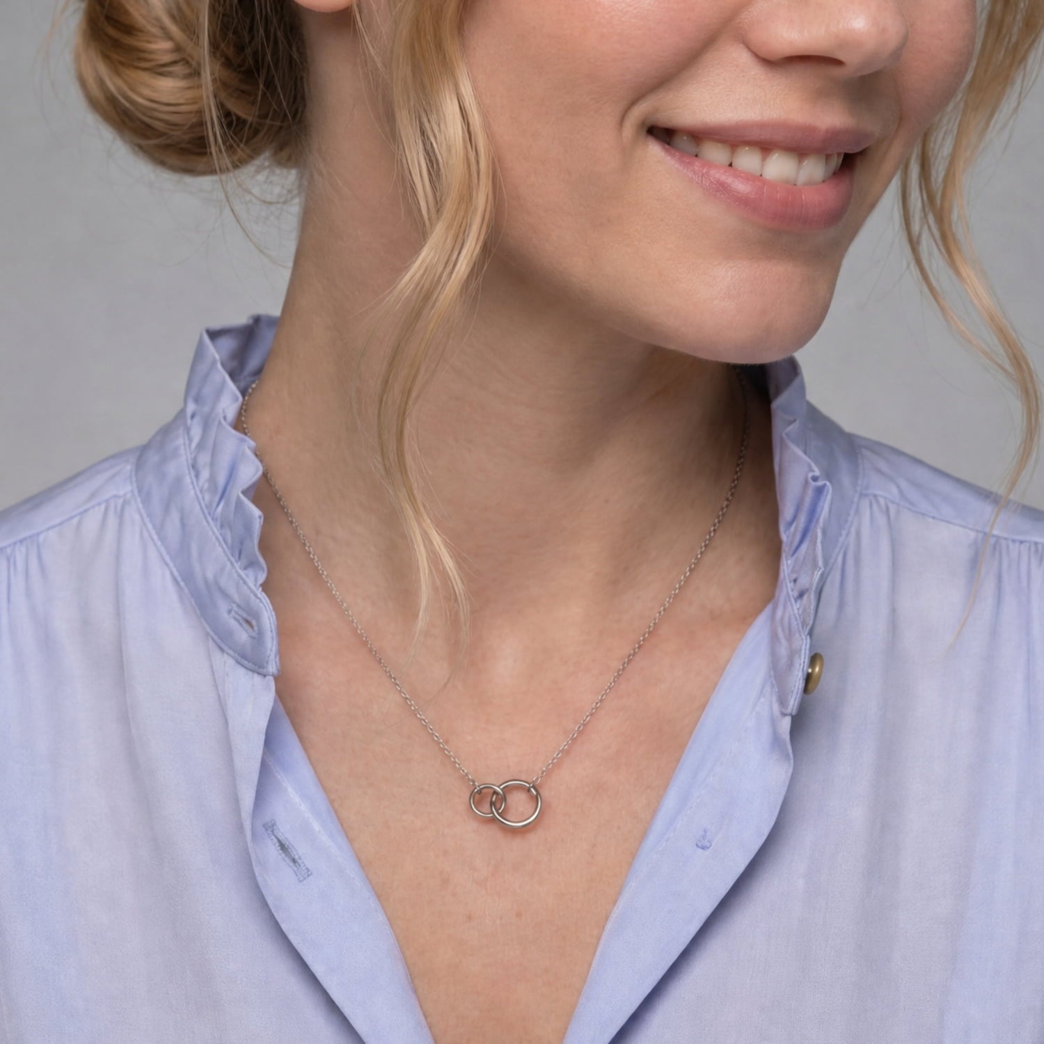 A close-up of a woman wearing a delicate silver chain necklace with  double interlocking rings, styled with a light blue button-down shirt.