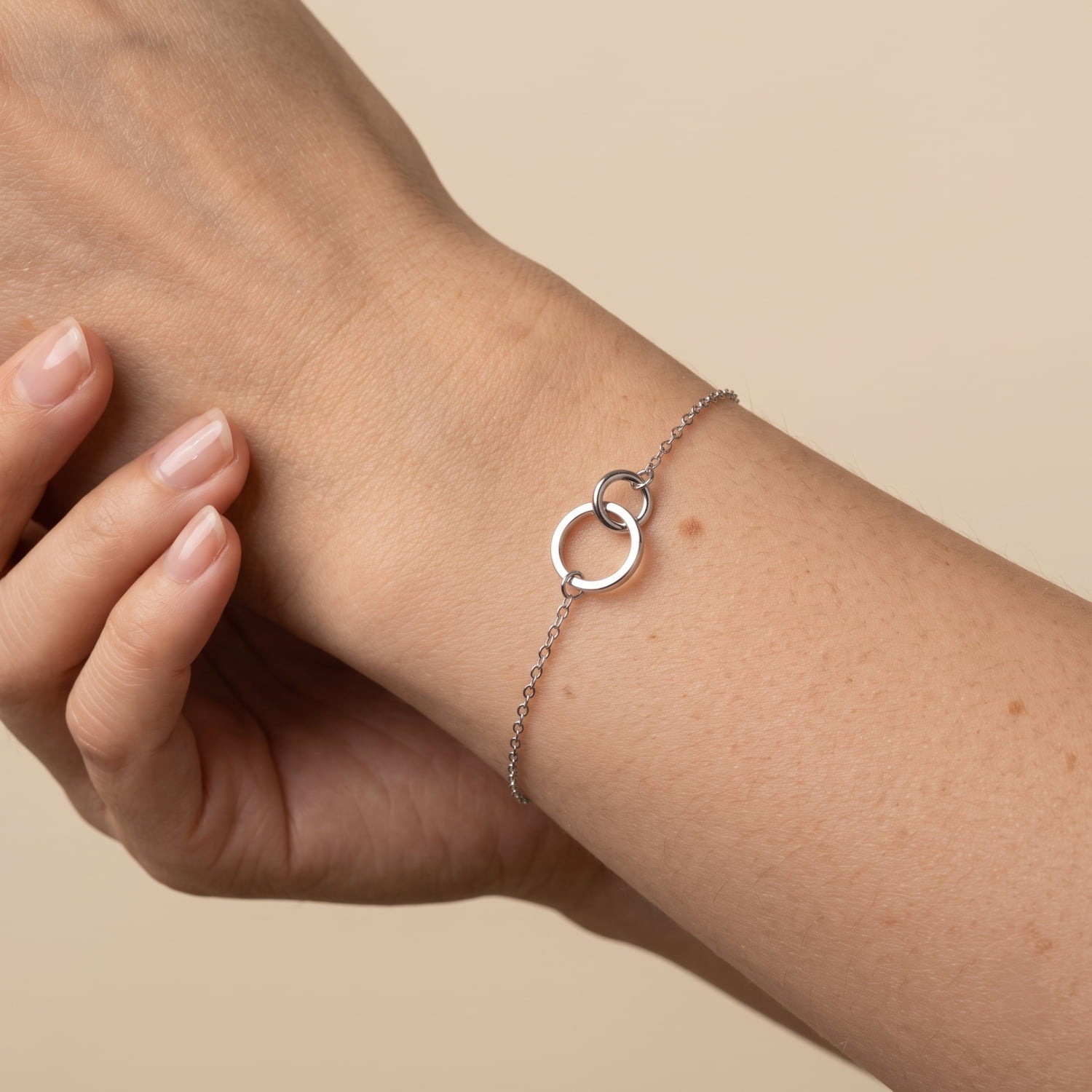 A delicate silver chain bracelet with two interlocking open circles, shown worn on a person's wrist against a soft beige background.