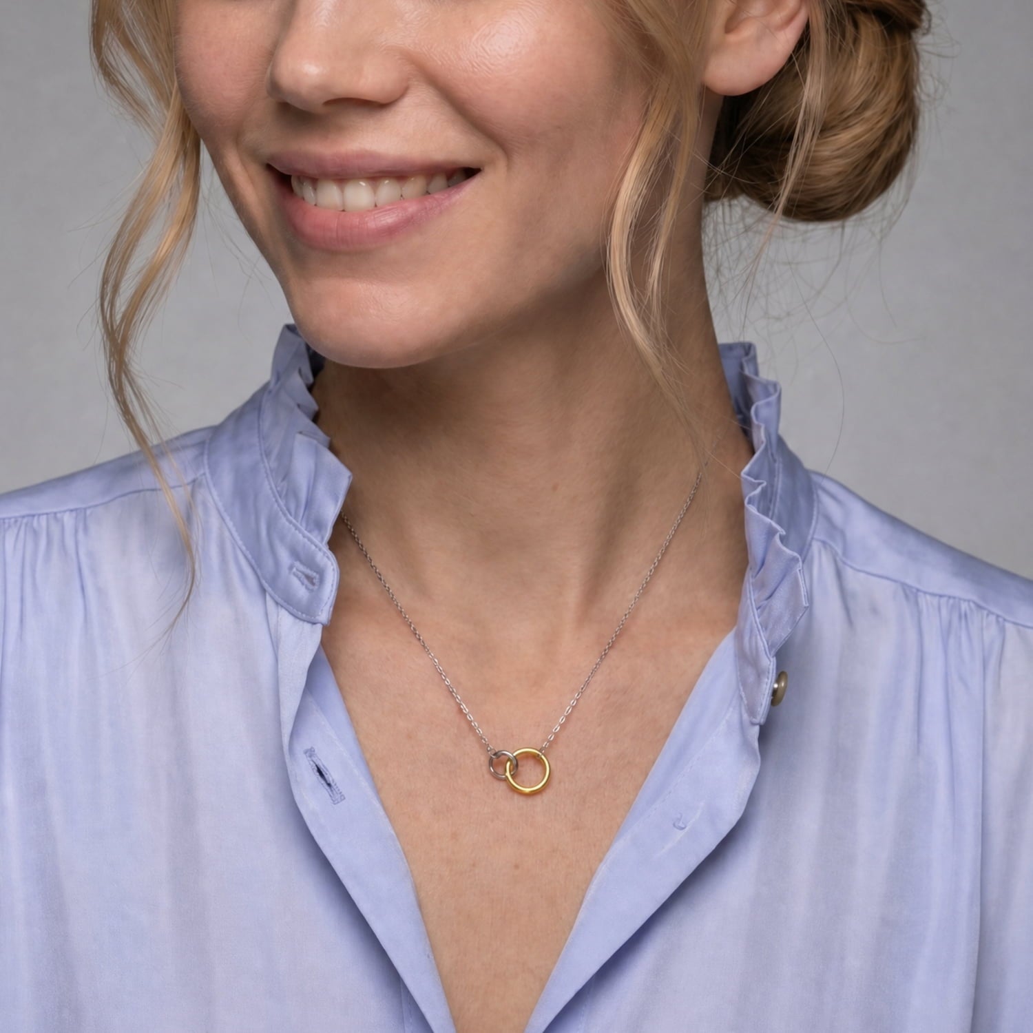A close-up of a woman wearing a delicate silver chain necklace with interlocking circles, paired with a light blue ruffled blouse against a neutral gray background.