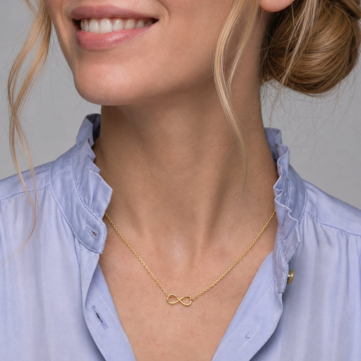 A close-up of a woman wearing a delicate gold vermeil infinity necklace with a small infinity symbol pendant, styled with a light blue ruffled-collar blouse.