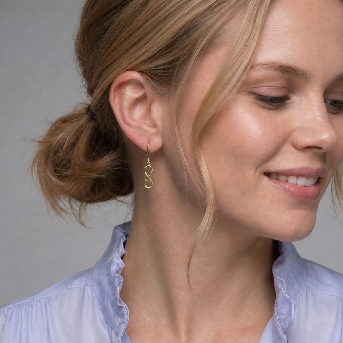 A close-up of a woman wearing a delicate gold vermeil infinity drop earring, with her hair in a low bun and a light blue blouse visible against a neutral gray background.