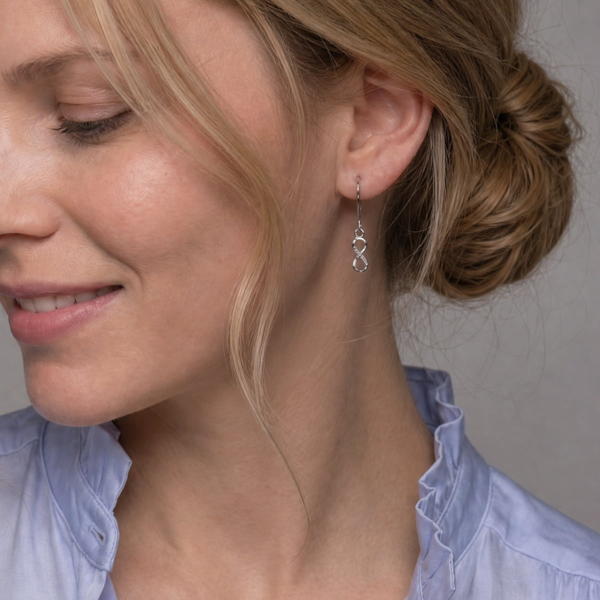A close-up of a woman wearing a delicate silver infinity drop earring set with lab grown diamonds, paired with a light blue ruffled blouse.