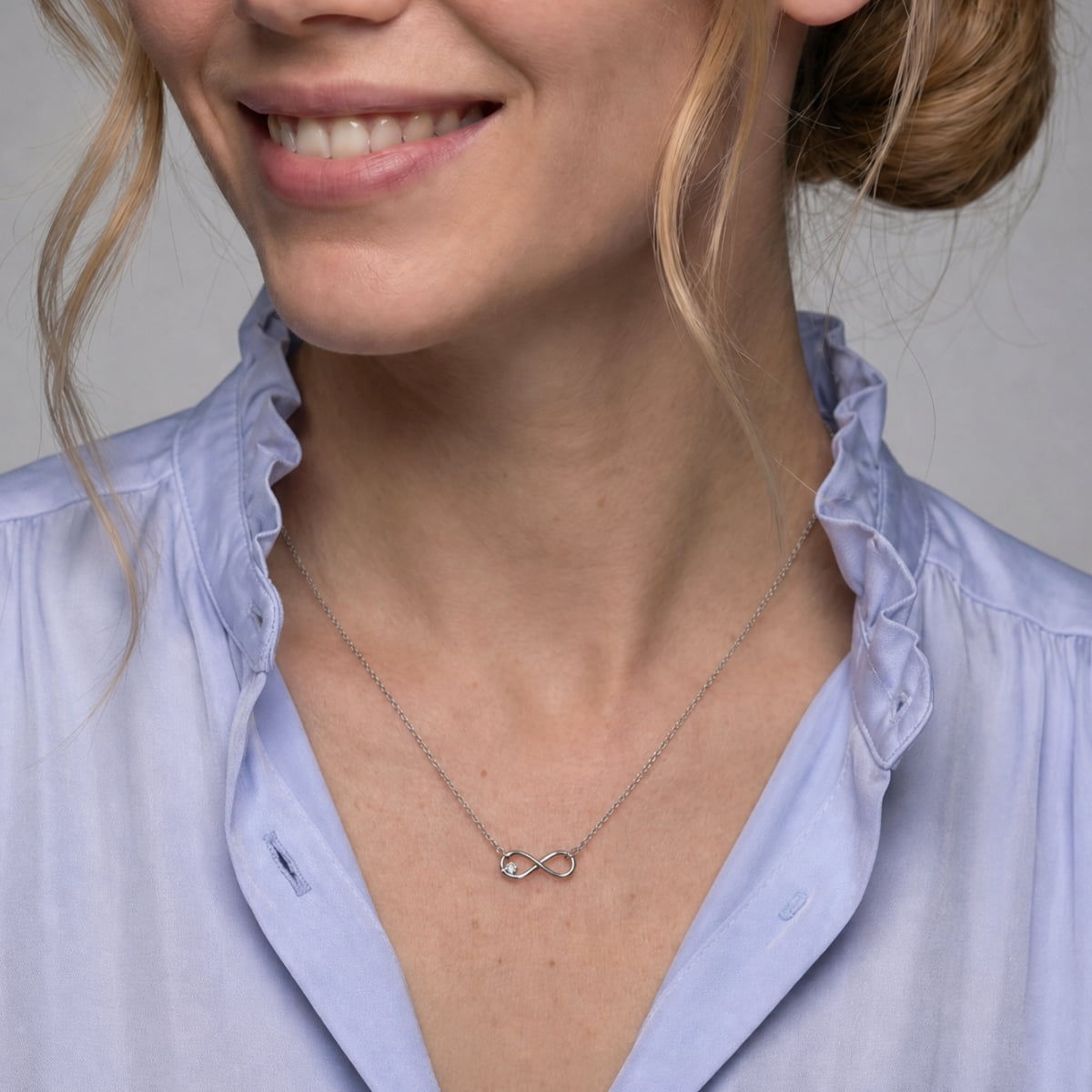 A close-up of a woman wearing a delicate silver infinity necklace with a small lab grown diamond, styled with a light blue ruffled blouse.
