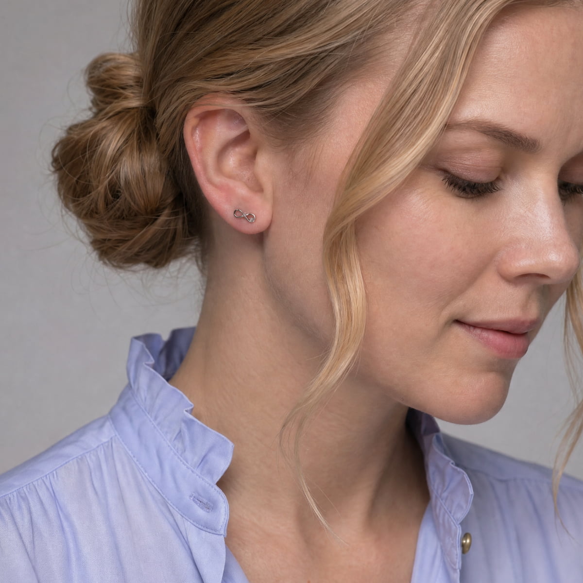A close-up of a woman wearing a tiny infinity and lab grown diamond earring on her ear, with her blonde hair in a bun and a light blue blouse against a neutral gray background.