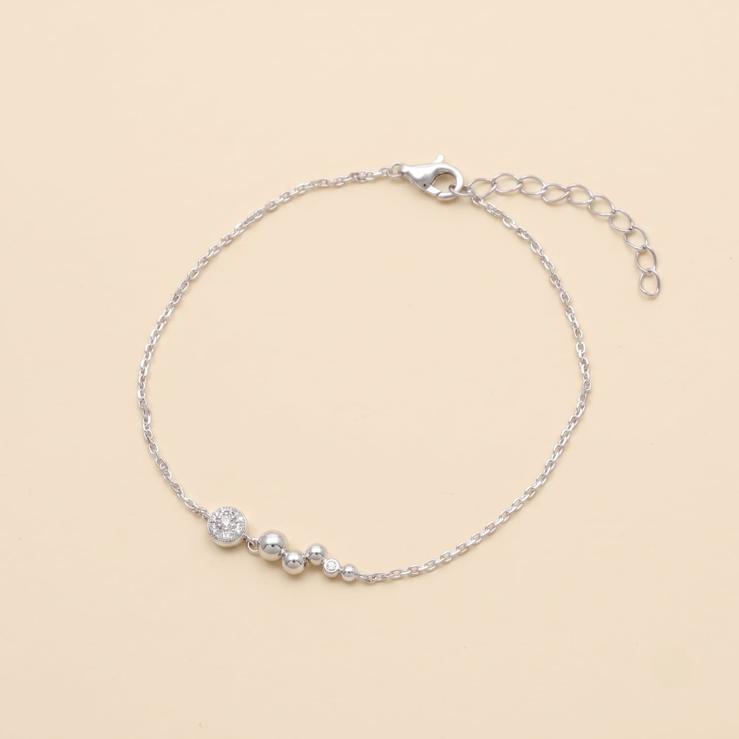A delicate silver chain bracelet with round bezel-set cubic zirconia and small polished silver beads, displayed on a light beige background.