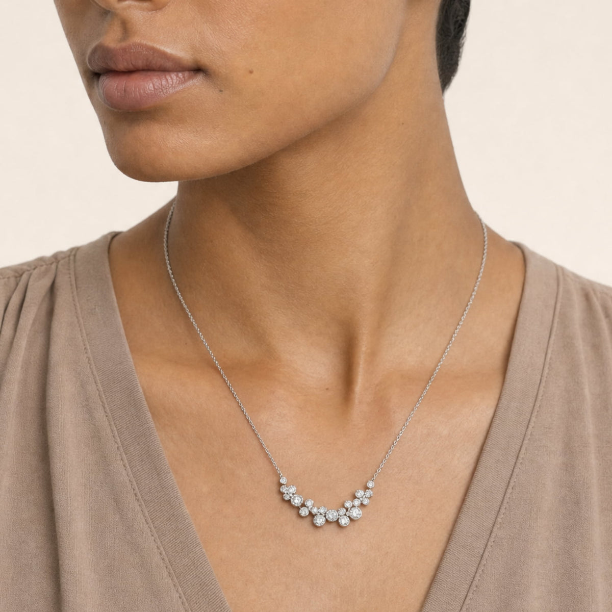 A close-up of a model wearing a silver necklace with a cluster of clear round cubic zirconia stones at the center, styled with a taupe brown top against a light beige background.