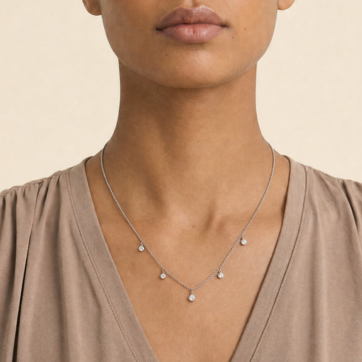 A close-up of a model wearing a delicate silver necklace with five small clear stones, paired with a taupe-brown V-neck top against a warm beige background.