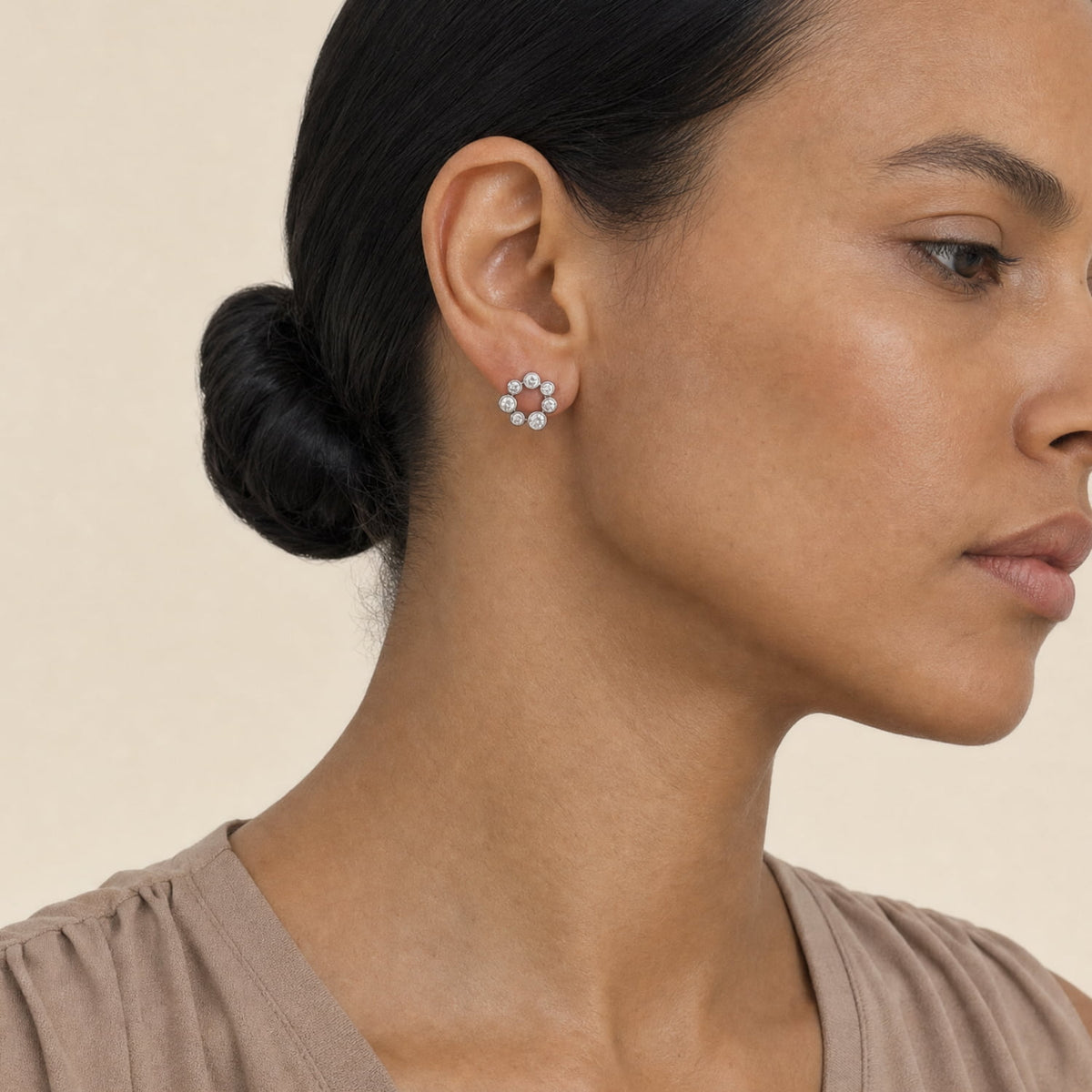 A close-up side profile of a woman with dark hair in a bun, wearing a silver stud earring with a halo of clear stones, against a warm beige background.