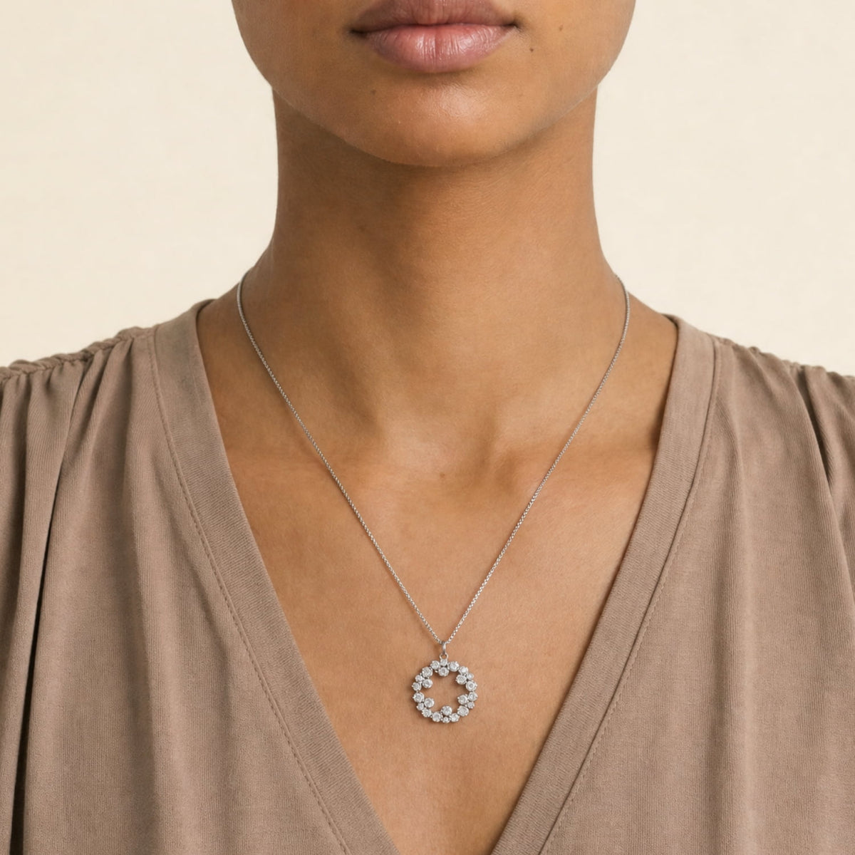 A close-up of a model wearing a silver chain necklace with a circular pendant encrusted with small clear stones, paired with a taupe brown V-neck top against a light beige background.