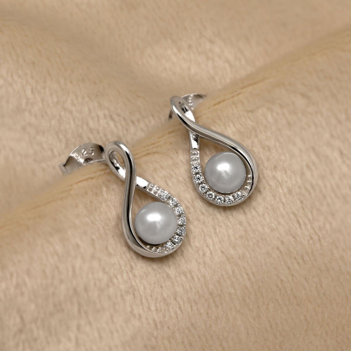 Silver infinity earrings with freshwater pearls and lab grown diamonds on a beige fabric background