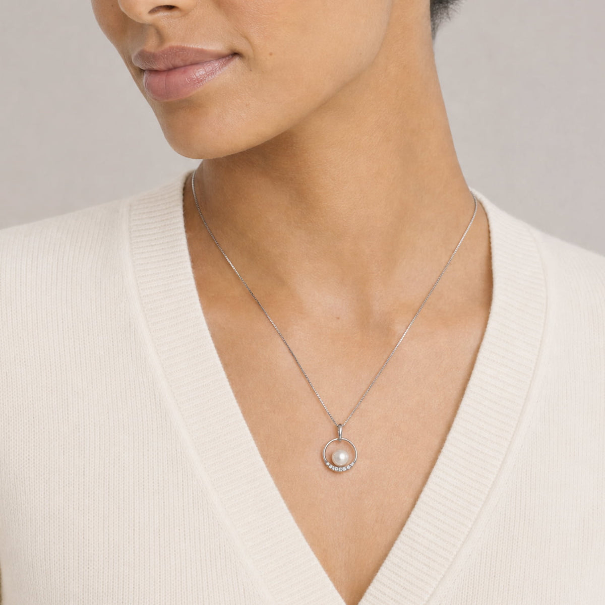 A close-up of a model wearing a delicate silver chain necklace with a circular pendant featuring a single pearl surrounded by small lab grown diamonds, paired with a cream-colored ribbed V-neck sweater.