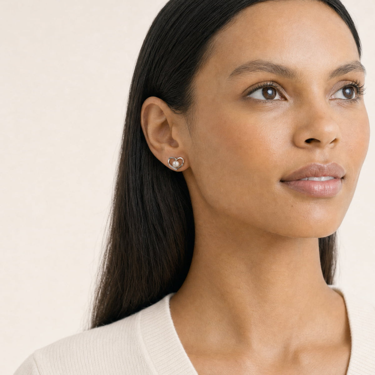 A close-up of a woman with long dark hair wearing a small heart-shaped silver stud earring with a freshwater pearl and lab grown diamonds, set against a soft beige background.